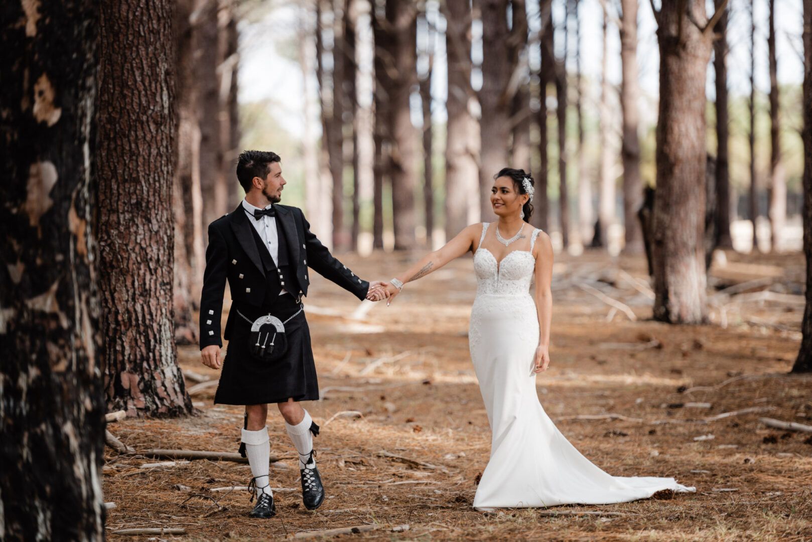 The groom and bride walk while holding hands in a pine forest in Wanneroo Pines, WA.