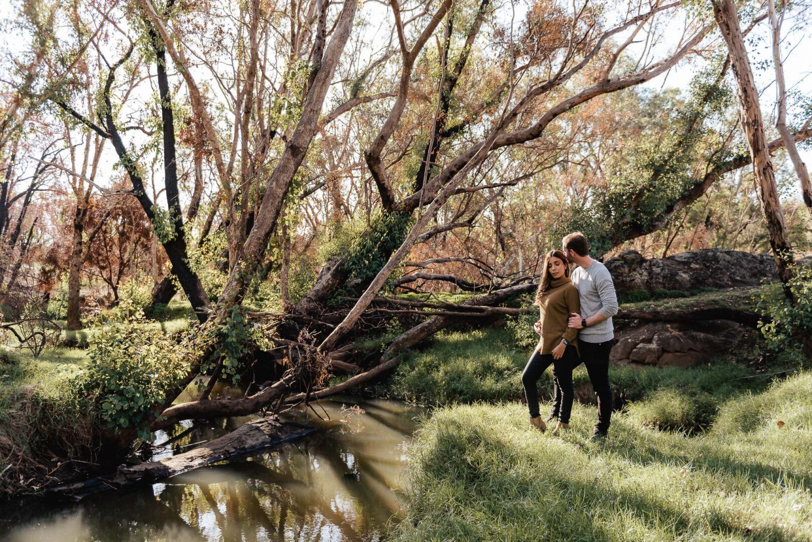 Two lovers stand together under a tree next to a river